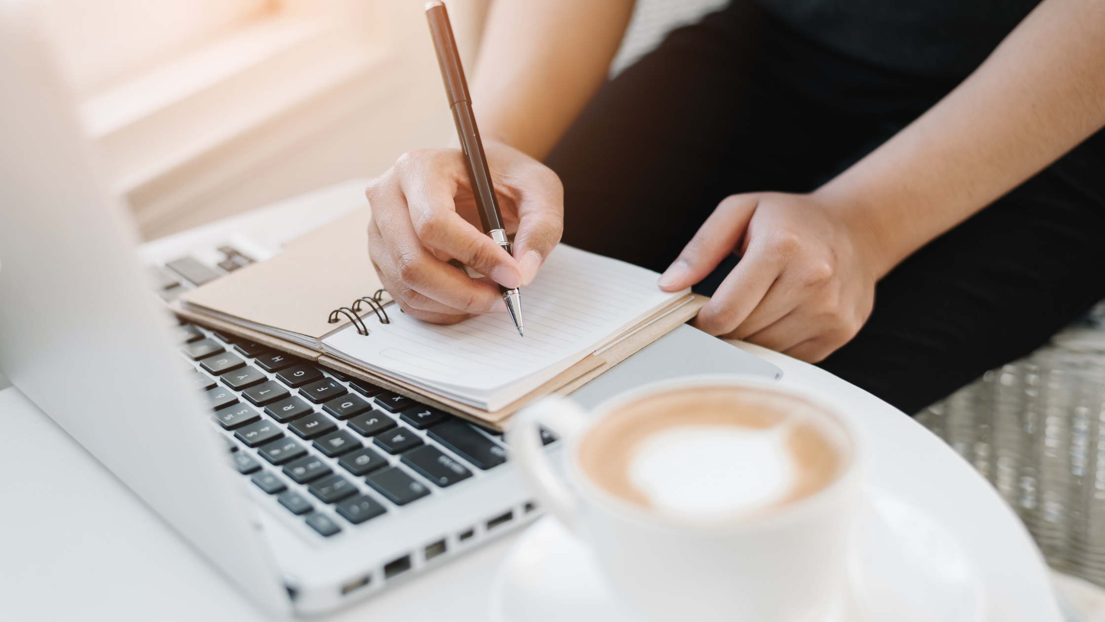 Image of lady writing in notebook, beside laptop, with coffee
